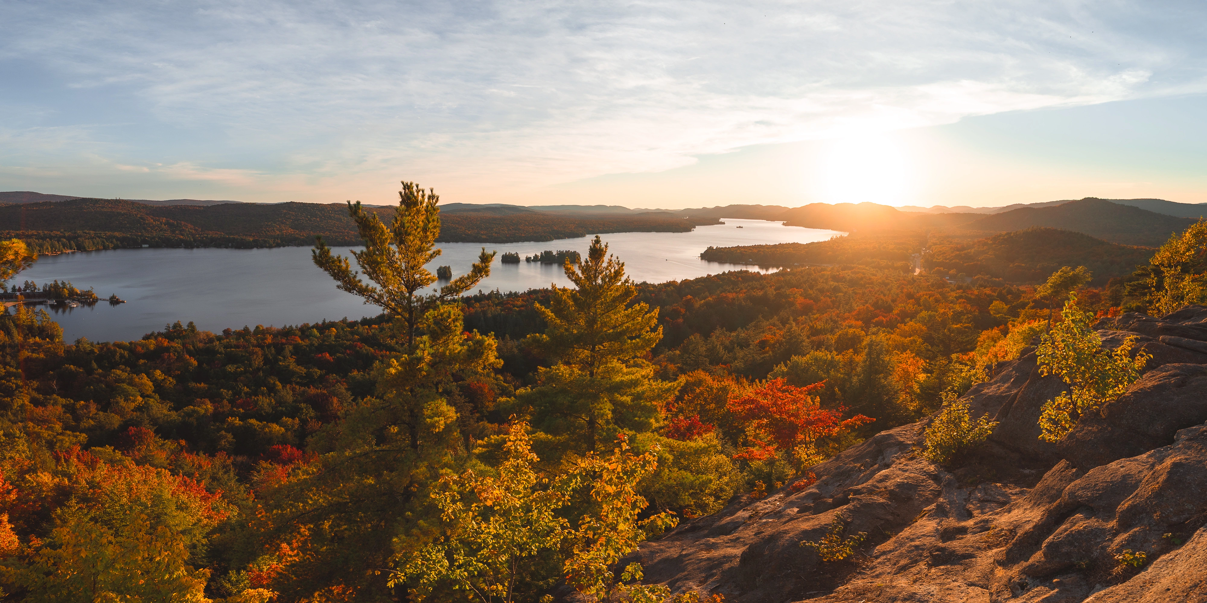 From Rocky Mountain near Inlet, New York, the sun drops toward Fourth Lake in the Fulton Chain of Lakes as the forest turns amber below me.