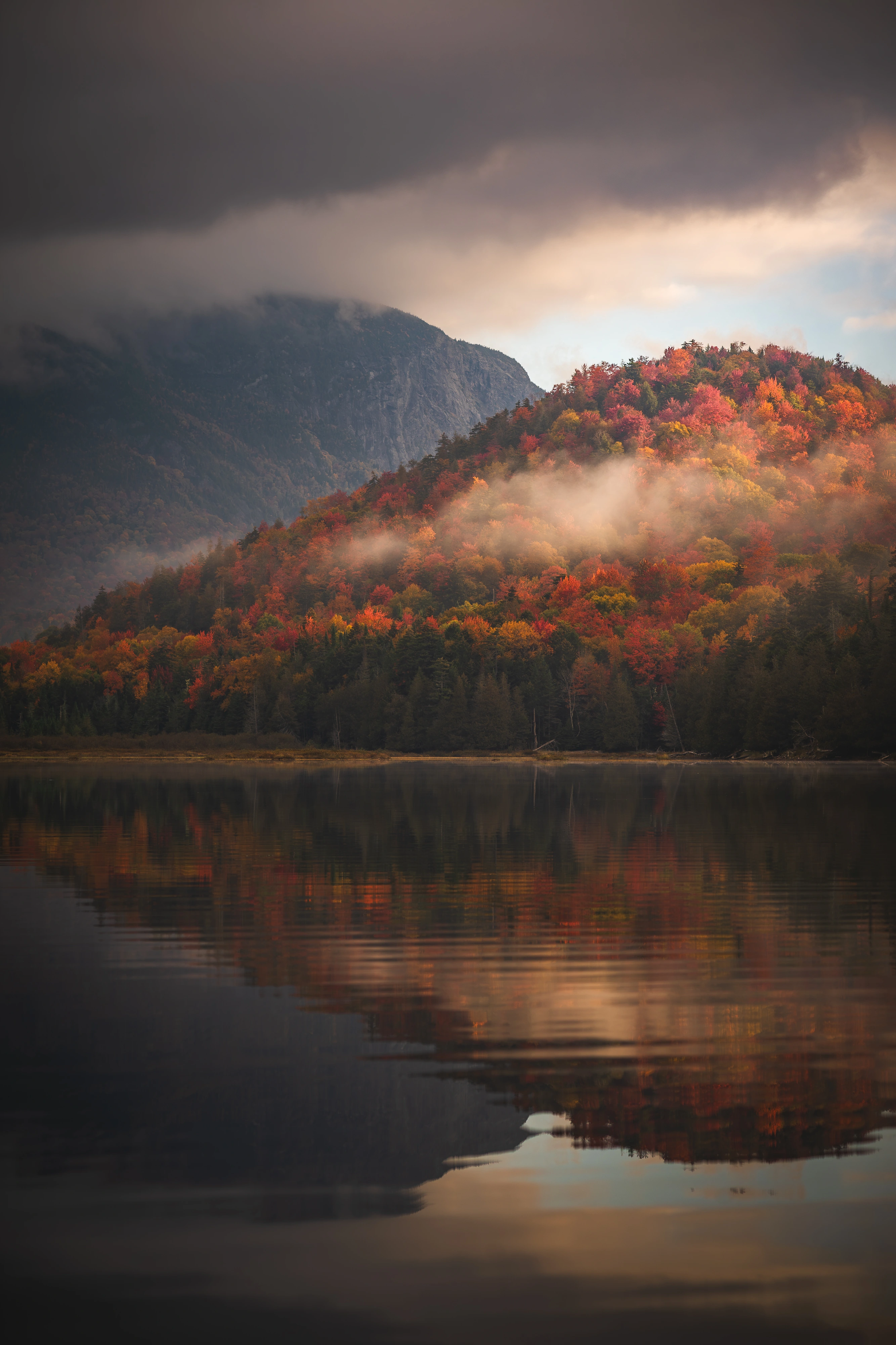 Still water mirrors the autumn ridge below Wallface as a band of fog crosses the trees. Henderson Lake, Adirondack High Peaks, New York.